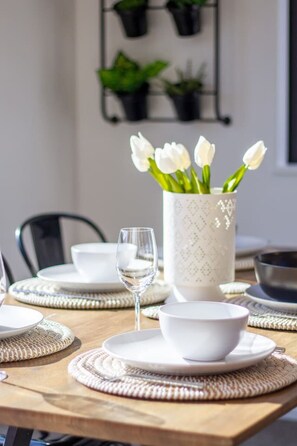 Una mesa de comedor dispuesta con platos, tazones y un vaso de agua, con un jarrón de tulipanes blancos en el centro.