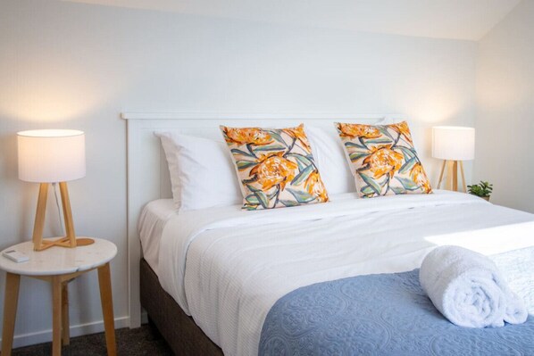 Una cama bien hecha con ropa blanca y dos almohadas decorativas con estampado floral. Hay dos mesitas de noche con lámparas, y una toalla enrollada sobre la cama.