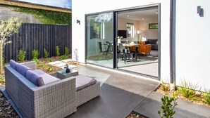 Un patio moderno al aire libre con un sofá de mimbre, una mesita y un vidrio que da acceso a un espacio interior con una mesa de comedor y sillas.