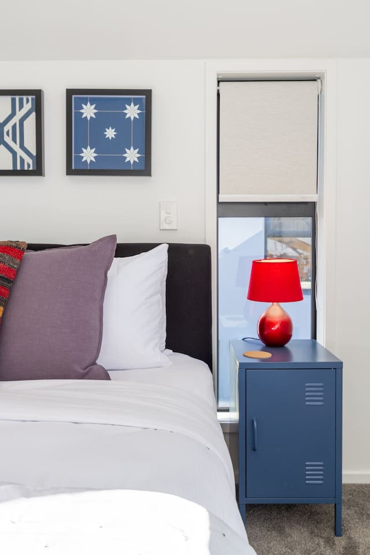 A bedroom with a bed, a nightstand with a red lamp, a blue cabinet, and two framed artworks on the wall.