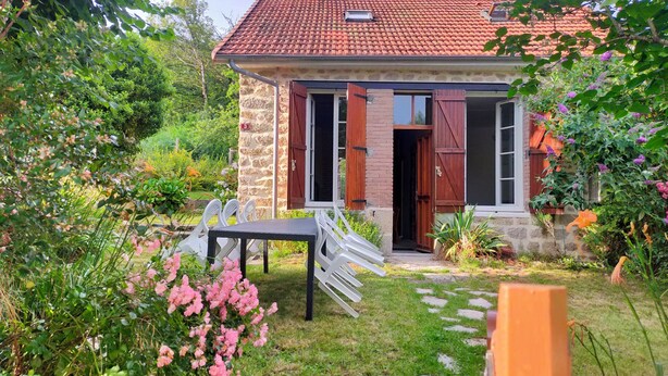 Une maison en pierre avec un toit recouvert de tuiles rouges, une porte en bois et un petit jardin agrémenté de fleurs roses et d’une chaise blanche.
