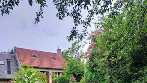 Une maison avec un toit recouvert de tuiles rouges, un mur en pierre et un jardin comprenant une pelouse et diverses plantes.