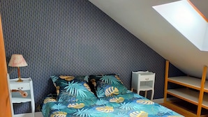 Une chambre avec un plafond en pente, un lit recouvert d’une couvre-lit orné de motifs de feuilles tropicales, une table de chevet avec une lampe et une petite commode blanche.