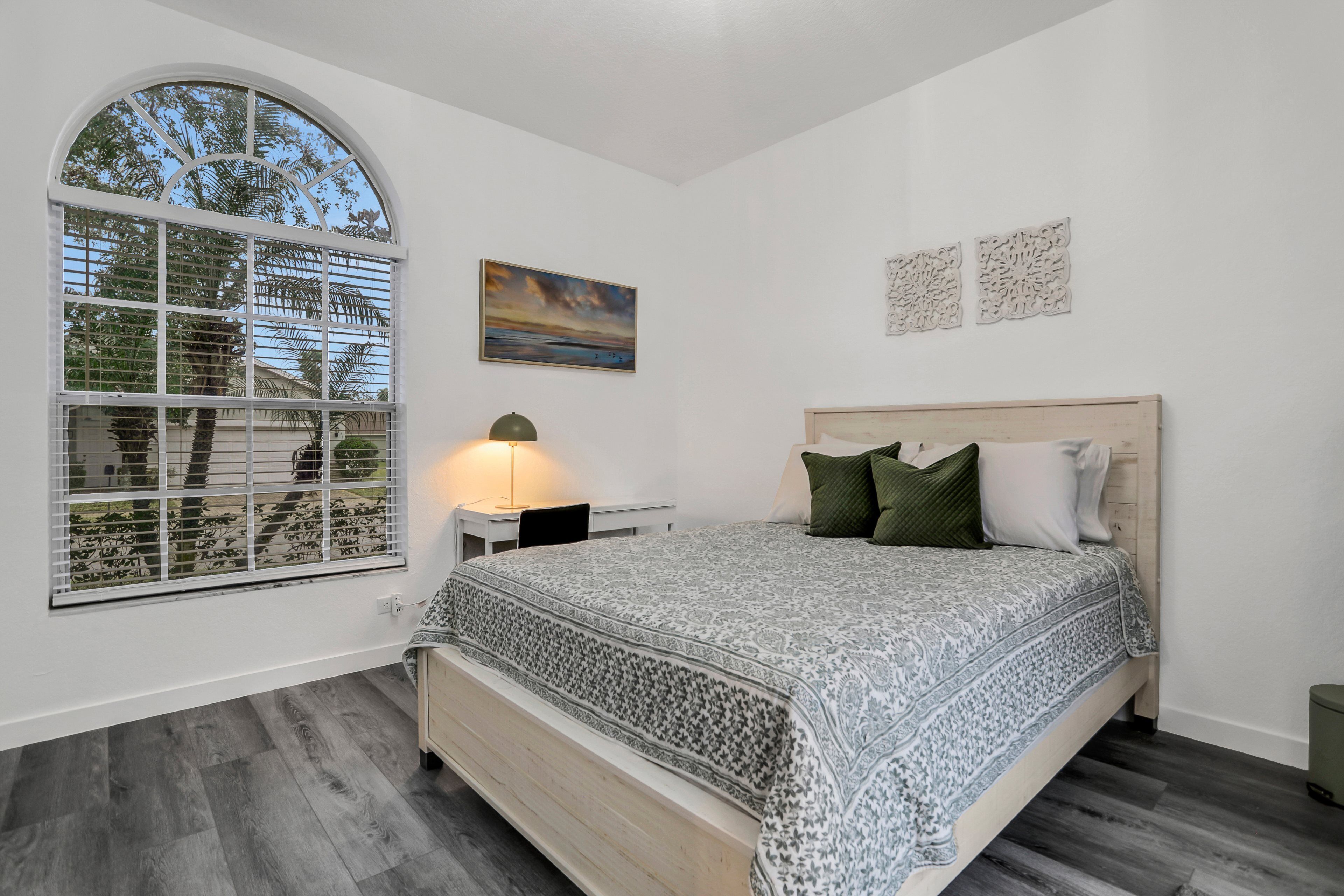 A bedroom with a large arched window, a bed with patterned bedding, a bedside table with a lamp, and a painting on the wall.