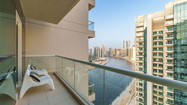 A balcony with white chairs and a glass railing overlooking a river and high-rise buildings. - Maison Privee - Superior Apt in Business Bay w/ Dubai Canal Views (Dubai)