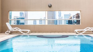 A swimming pool with two white lounge chairs, a view of a cityscape through a large window, and a beige wall. - Maison Privee - Superior Apt in Business Bay w/ Dubai Canal Views (Dubai)