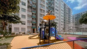 A playground with slides and climbing structures, surrounded by a fence, with residential buildings in the background.
