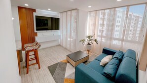 A modern living room with a blue sofa, a wooden coffee table, a TV mounted on the wall, and a window with a view of high-rise buildings.