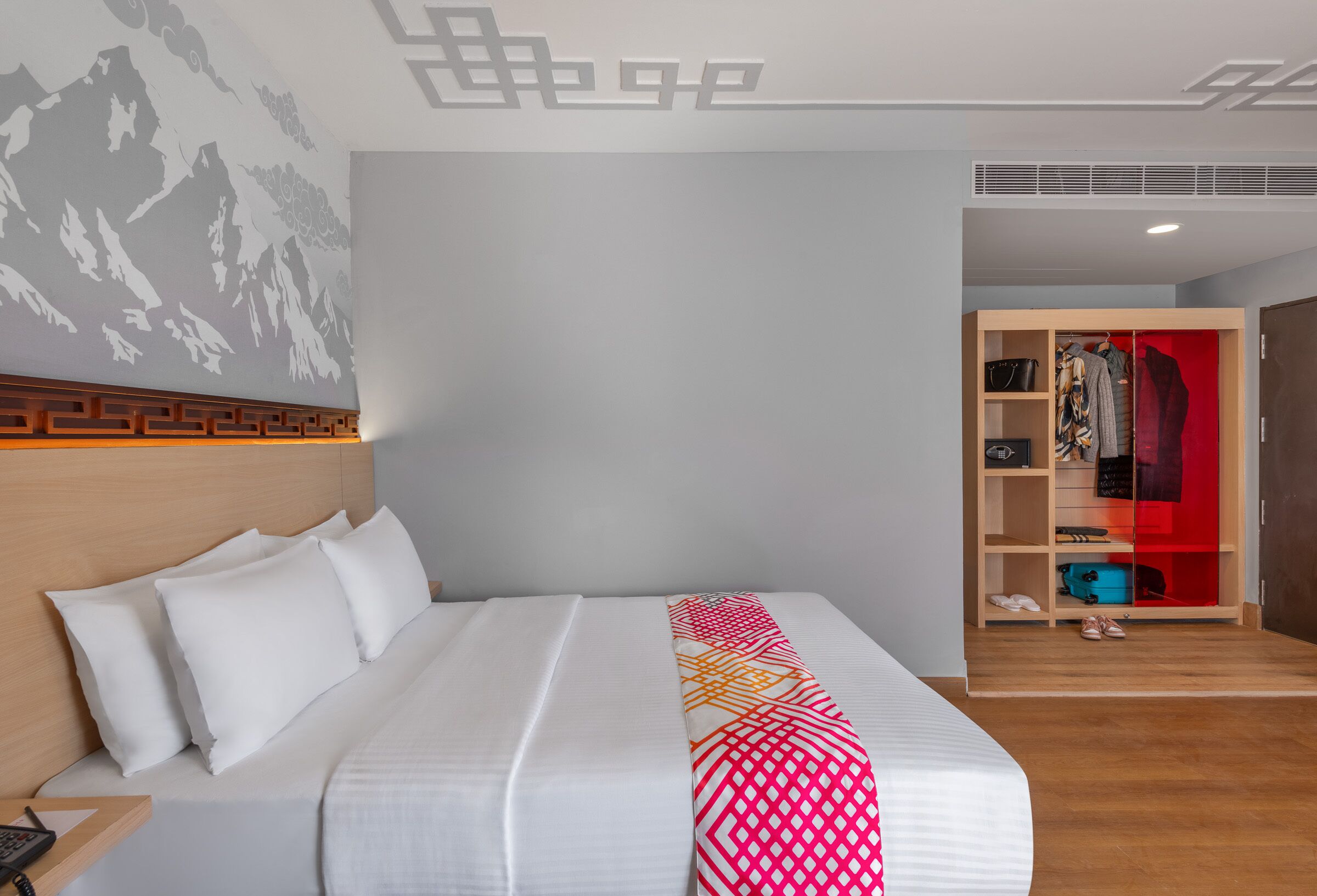 A hotel room with a bed, a wooden headboard, a wall-mounted phone, a closet with a red interior, and a patterned throw blanket.