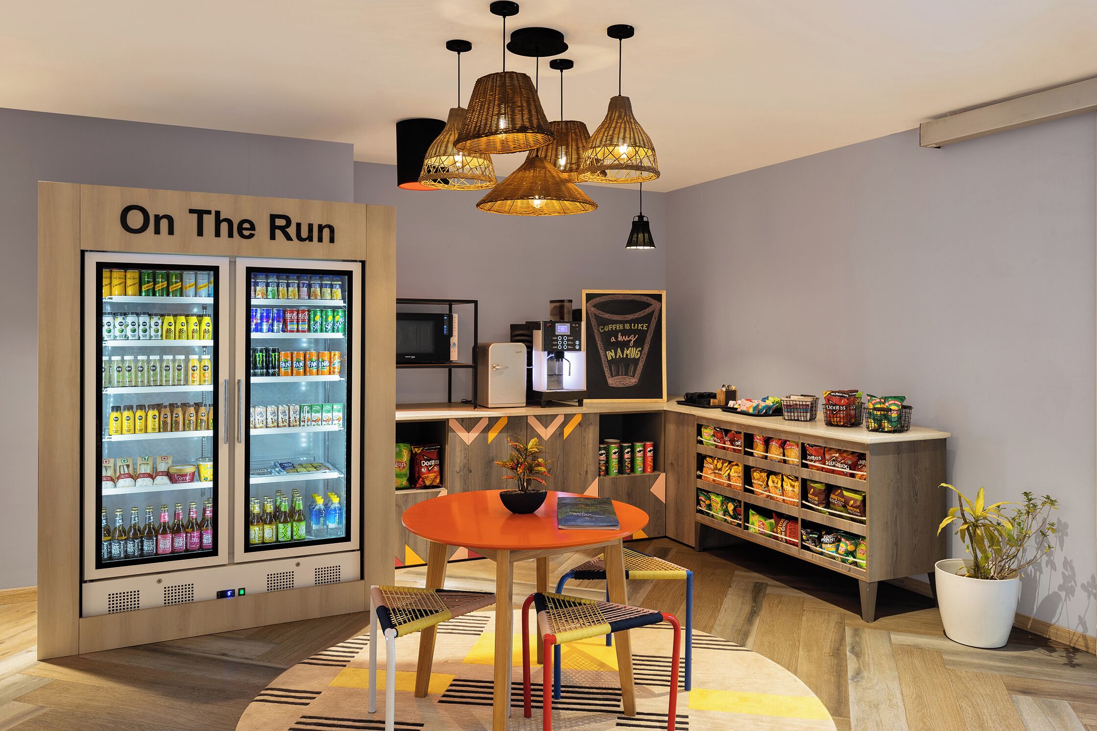 Une chambre moderne équipée d’un réfrigérateur « On The Run » rempli de boissons, d’une machine à café, d’une table à manger avec des chaises et d’une bibliothèque contenant des en-cas.