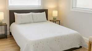 Une chambre à coucher comprenant un lit, deux tables de chevet, un ventilateur de plafond et deux fenêtres équipées de stores.