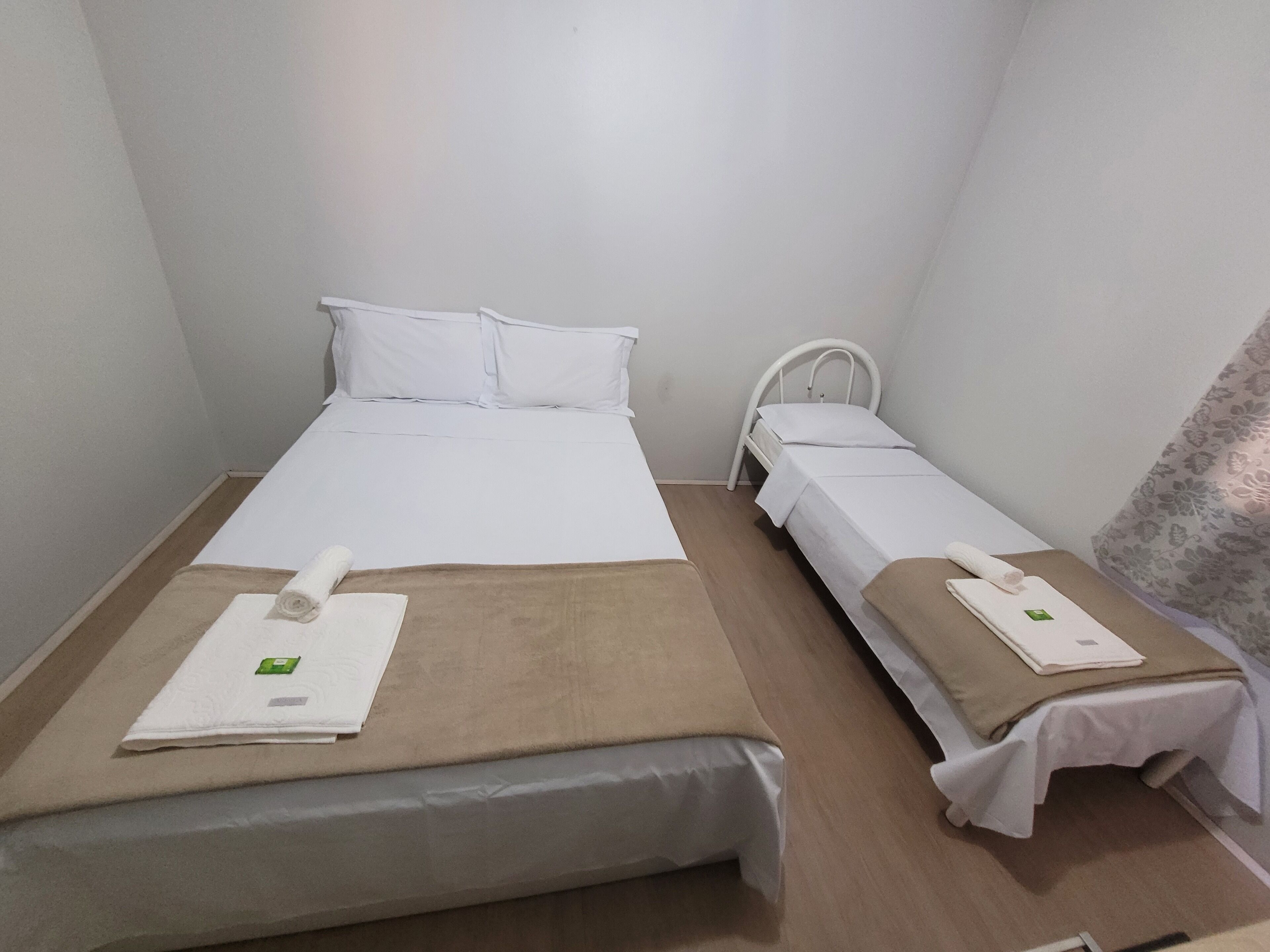 Quarto pequeno e limpo com duas camas, cada uma com colchão e almofadas brancas e um cobertor marrom. Há toalhas sobre as camas.