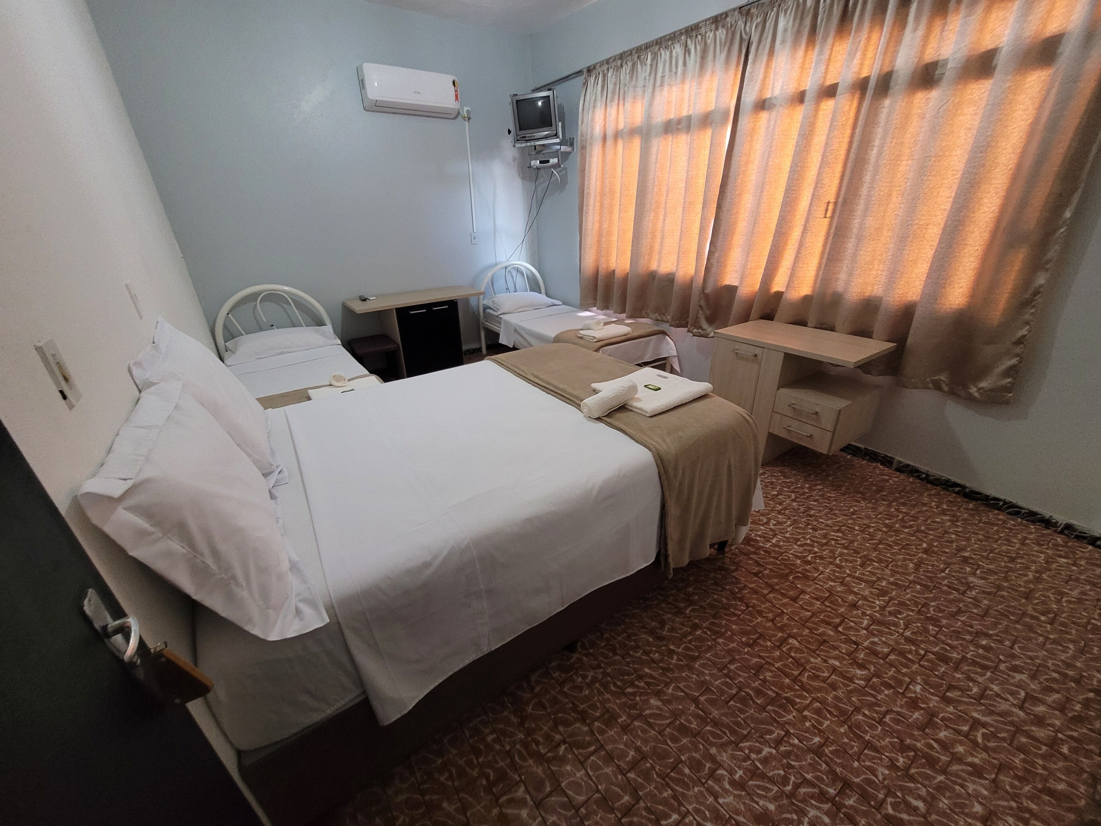 Quarto de hotel com duas camas, uma escrivaninha, uma cadeira, uma televisão e uma janela com cortinas.
