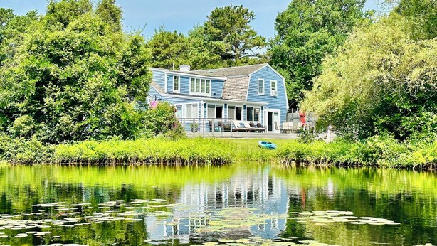 A two-story house with a porch, surrounded by lush greenery and a calm pond with lily pads. - Beautiful waterfront home- mins to beach, fishing /kayak right from the backyard (Yarmouth)