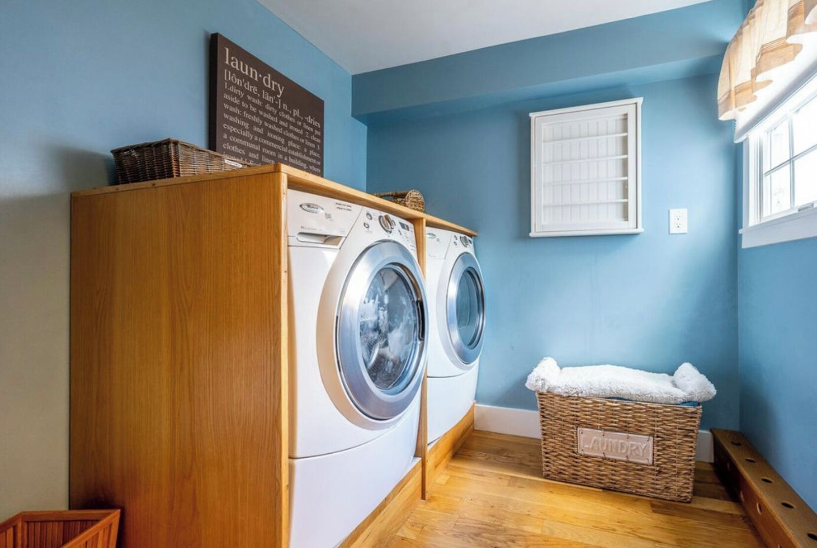 A laundry room with two front-loading washing machines, a wooden cabinet, a wicker laundry basket, and a sign with laundry-related instructions.