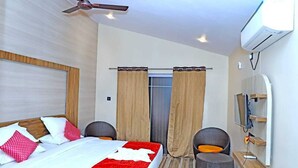 A hotel room with a bed, two chairs, a TV, and an air conditioning unit.
