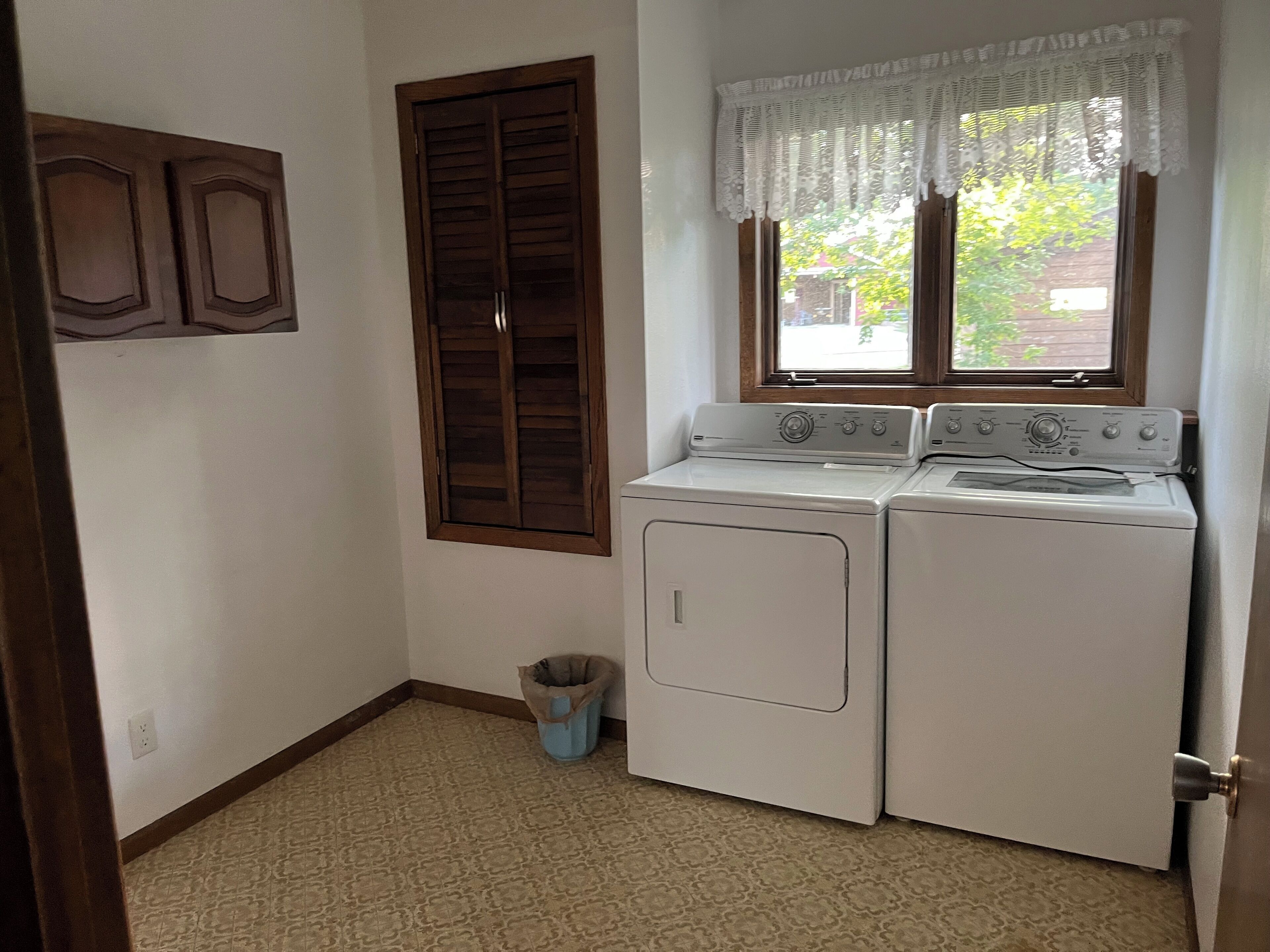 A laundry room with two stacked washers and dryers, a window with a lace curtain, a wooden door, and a trash can.