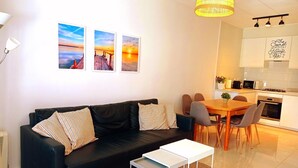 Un salon moderne avec un canapé noir, une petite table blanche et un coin repas comprenant une table et des chaises en bois. Trois tableaux encadrés ornent le mur, et on y trouve une cuisine avec des meubles blancs, ainsi qu’un luminaire suspendu.