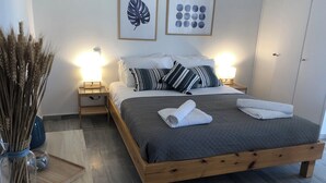A neatly arranged bedroom with a wooden bed, white pillows, and a grey quilted bedspread. There are two wall-mounted lamps, a framed leaf artwork, and a vase with reeds on the nightstand. - Zen Studio 2 min from the beach with a spacious veranda, free WiFi, and A/C. (Rethymno)