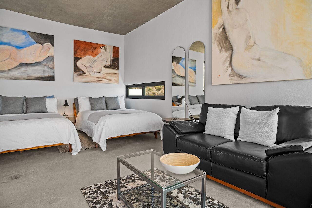 A modern bedroom with two beds, a black leather sofa, and abstract paintings on the walls.