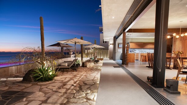 A modern outdoor patio with stone flooring, lounge chairs, and umbrellas, leading to a well-lit interior with wooden furniture and a kitchen area.