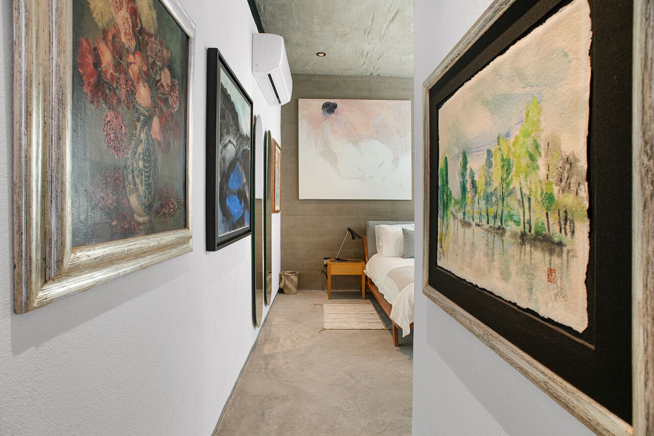 A hallway with framed artworks on the walls, a bed with a wooden frame, and a small table with a lamp.