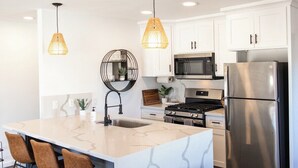 Une cuisine moderne avec des armoires blanches, un réfrigérateur en acier inoxydable, un four à micro-ondes et une cuisinière à gaz. On y trouve deux luminaires suspendus, un évier et un tabouret de bar.