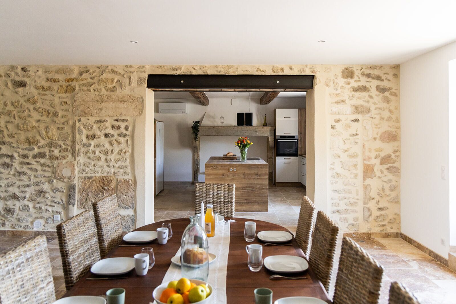 Un coin repas agrémenté d’un mur en pierre, d’une table en bois, de chaises en osier et offrant une vue sur une cuisine équipée d’appareils électroménagers modernes.