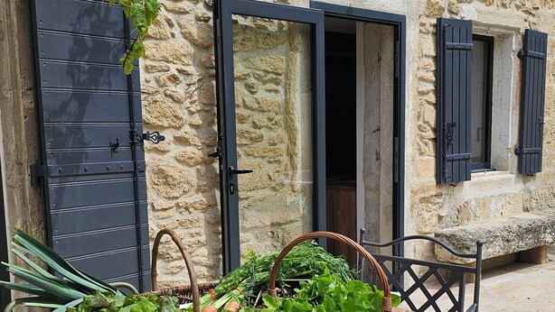 Un bâtiment en pierre avec une table en bois et des chaises en métal, des paniers de légumes et une vigne grimpante.
