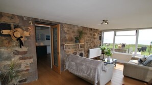 Ein gemütliches Wohnzimmer mit Steinwand, einer gepolsterten Couch, einem Couchtisch mit Pflanze und Blick ins Freie.