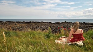 Eine Frau sitzt auf einer Decke an einem felsigen Ufer, im Hintergrund das Meer.