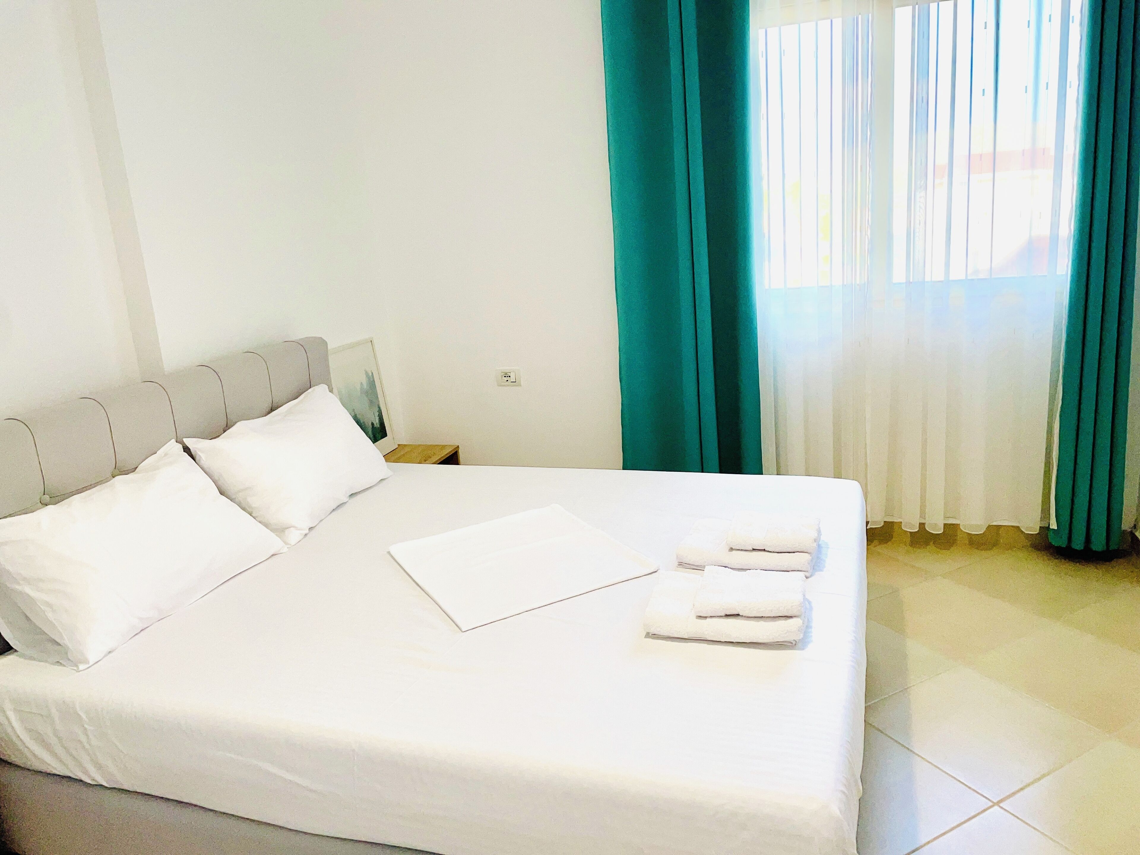 A neatly made bed with white linens and pillows, a small wooden nightstand, and a window with teal curtains.