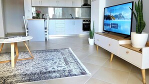 Eine moderne Küche mit weißen Schränken, ein Essbereich mit einem runden Tisch und ein Wohnzimmer mit Fernseher und Pflanzen.