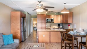 A modern kitchen with wooden cabinets, a central island, and a dining area with a table and chairs. - Wonderful South Maui Beach Condos  (Kihei)