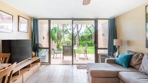 A living room with a sofa, TV, wooden furniture, and a sliding glass door leading to an outdoor patio with chairs and a table. - Wonderful South Maui Beach Condos  (Kihei)
