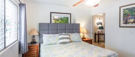 A bedroom with a bed, a ceiling fan, a nightstand with a lamp, a framed picture on the wall, and a window with curtains.