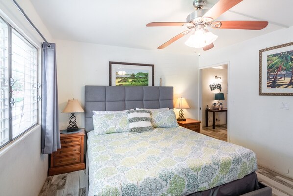 A bedroom with a bed, a ceiling fan, a nightstand with a lamp, a framed picture on the wall, and a window with curtains.