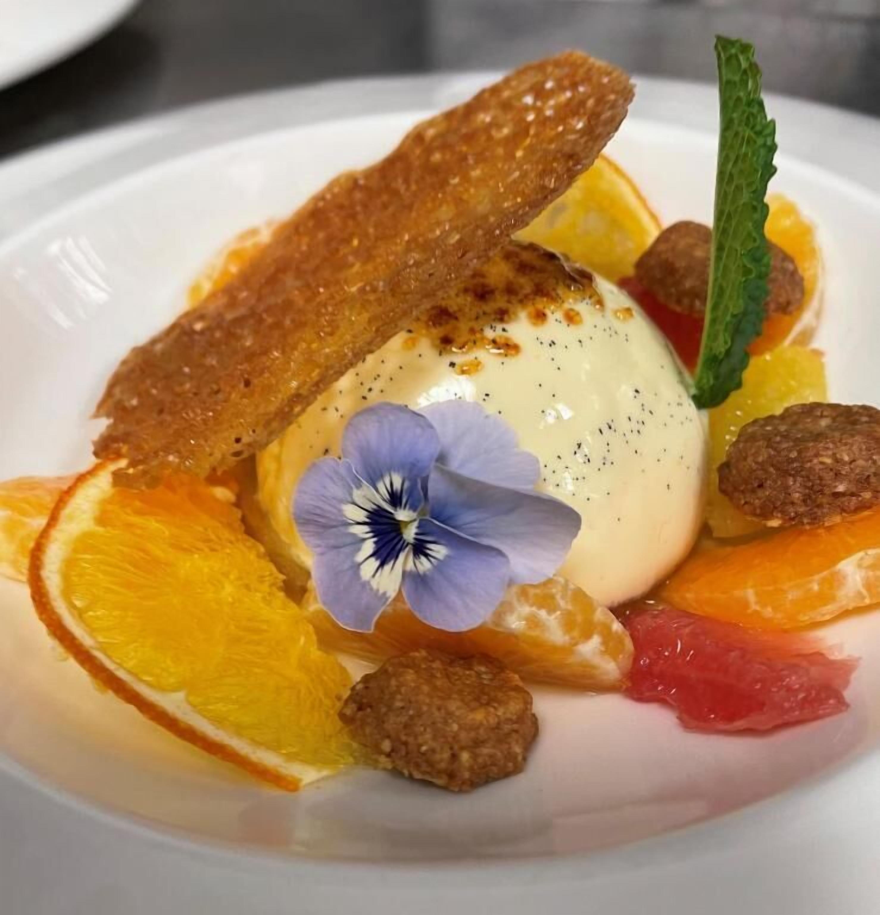 Un postre con una bola de helado, rodajas de naranja, una flor y nueces en un plato.