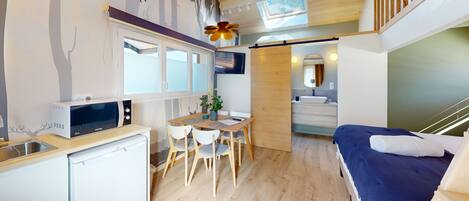 Cocina compacta con microondas, lavamanos y refrigerador pequeño. Hay una zona de comedor con mesa y sillas, y una cama con manta azul.
