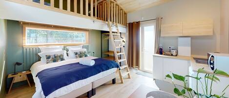 A bedroom with a lofted sleeping area, a bed with a blue and white duvet, a wooden ladder, a small desk with a kettle, and a potted plant.