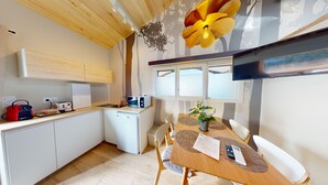 Eine kleine Küche mit weißen Schränken, einem kleinen Kühlschrank und einer Mikrowelle. Ein Esstisch mit Stühlen und ein Blumentopf sind ebenfalls vorhanden.