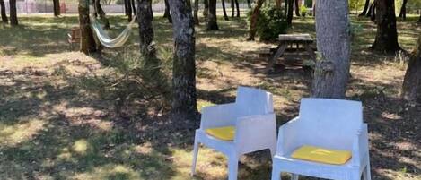 Two white plastic chairs with yellow cushions in a shaded outdoor area with trees.