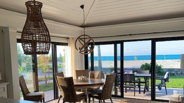 Cozinha moderna com uma ilha grande, área de jantar com cadeiras de vime e vista para a praia através de portas deslizantes amplas.