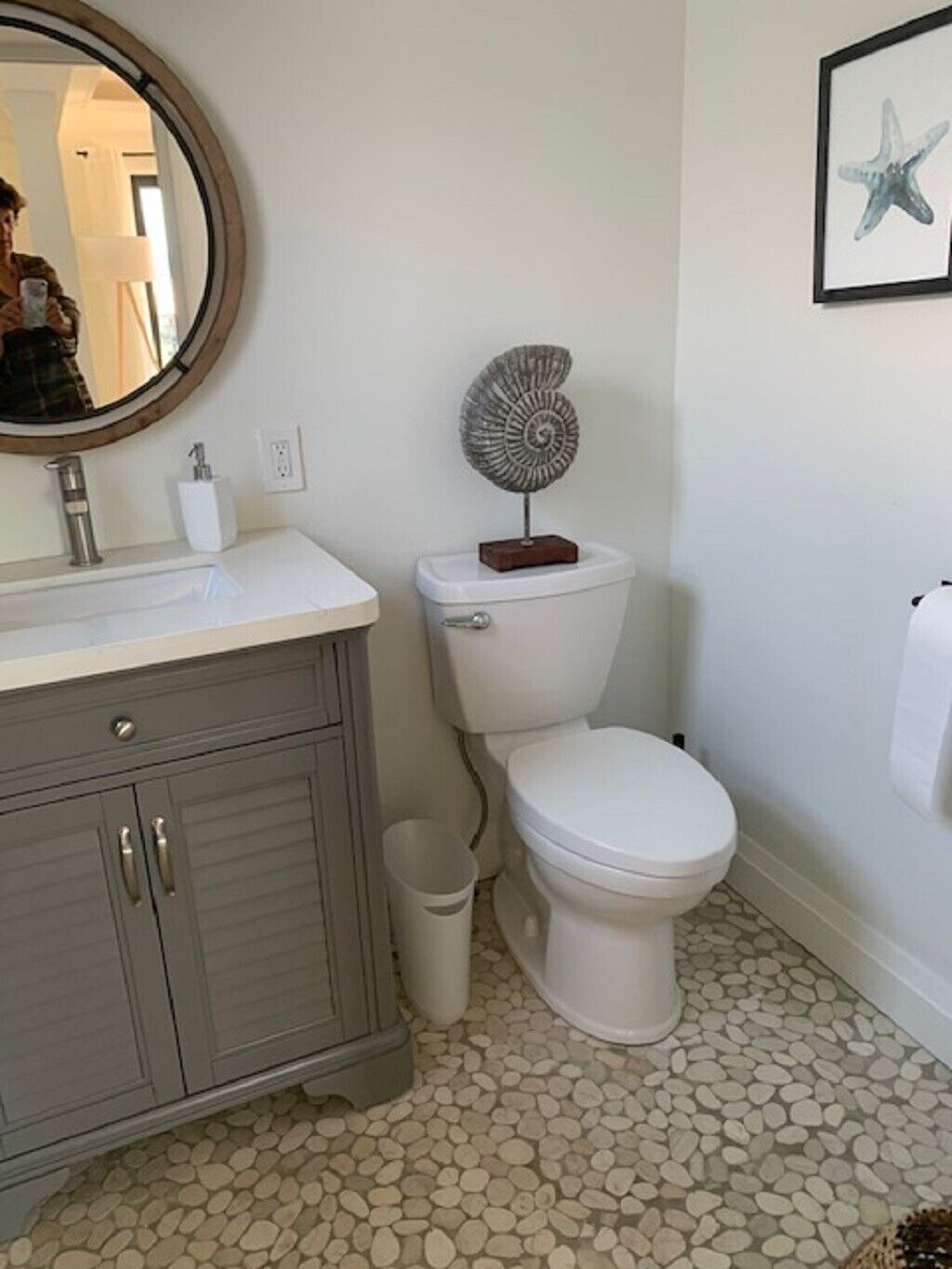 A bathroom with a white toilet, a grey cabinet, a round mirror, a starfish decoration, and a pebble-patterned floor.