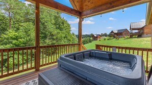 Un jacuzzi en una terraza de madera con vistas a un paisaje verde y una cabaña de madera al fondo.