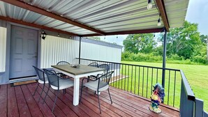 Une terrasse extérieure couverte, avec une table et des chaises, une porte et une vue sur une cour verdoyante.