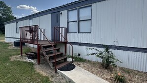 Une maison mobile de plain-pied, dotée d’un porche à l’avant, d’un petit jardin et offrant une vue sur un ciel dégagé.