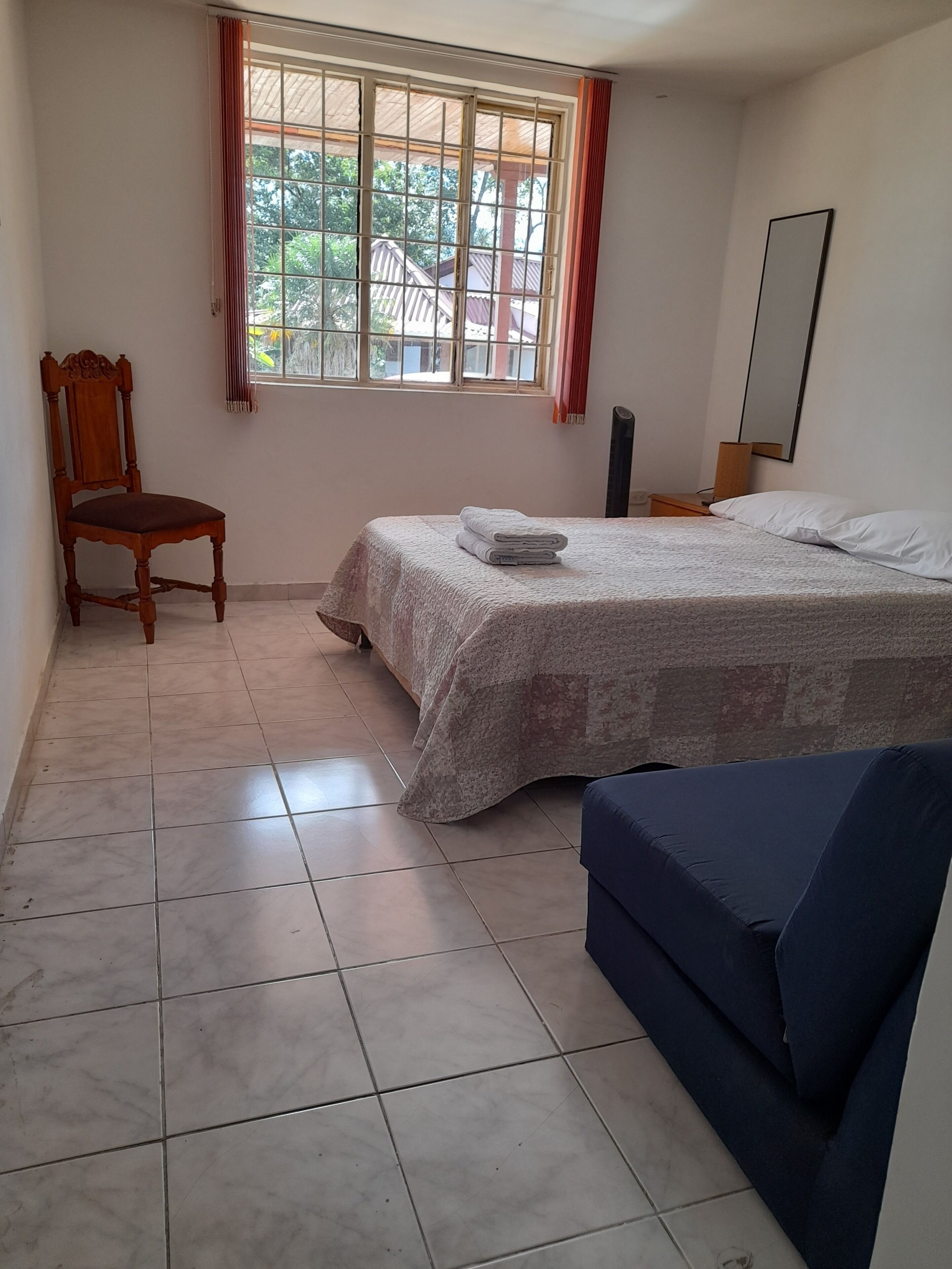 Un dormitorio con cama, una silla, una mesita de noche y una ventana con cortinas.