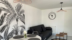 A modern living room with a black sofa, a round dining table with placemats, a large wall mural of leaves, and two pendant lights. - Studio Cosy (Mende)