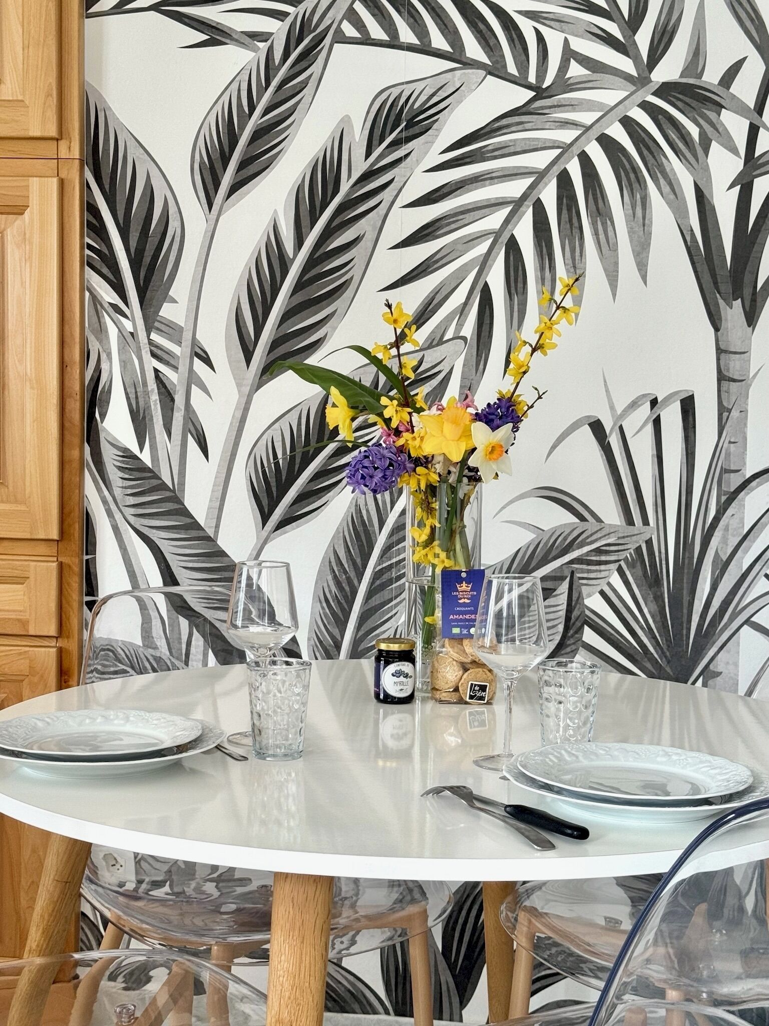 A round dining table with clear glassware, a vase with flowers, and a jar of jam.